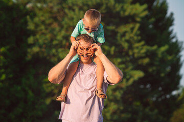 Happy father and son spending time together and having fun outside. Small boy sitting on shoulders of his father and playing with him. Father laughs with closed eyes.