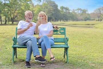 senior caucasian grandfather and grandmother sitting in park while using video call from smart phone to grandchildren with happy face and laughing