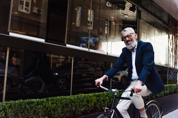 happy middle aged man in glasses and blazer riding bicycle on modern urban street