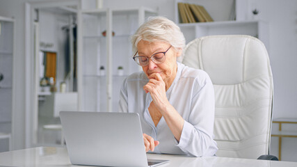 Tired sleepy old lady manager types on grey laptop at white table sitting on large chair against different shelves