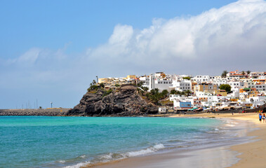 View of part of the town of Morro Jable with white buildings on a rocky shore and the beach with turquoise blue ocean water. Fuerteventra, Canary Islands, Spain