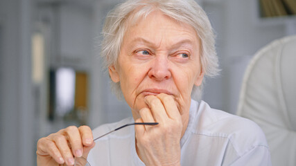 Thoughtful senior businesswoman with grey hair holds glasses and touches chin sitting on large...