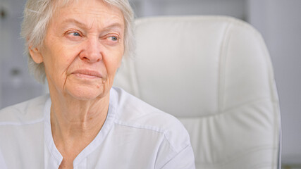 Fototapeta na wymiar Old wise grey haired mother with blue eyes looks aside with sad face sitting in white chair under bright light