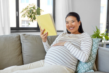 pregnancy, people and leisure concept - happy smiling pregnant asian woman sitting on sofa and reading book at home