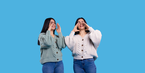 Brunette sisters screaming with their arms keeping hands near mouth standing on a blue background.