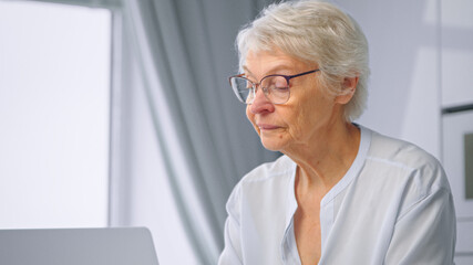 Upset aged businesswoman with grey hairstyle wipes nose with paper napkin and types on laptop sitting at home