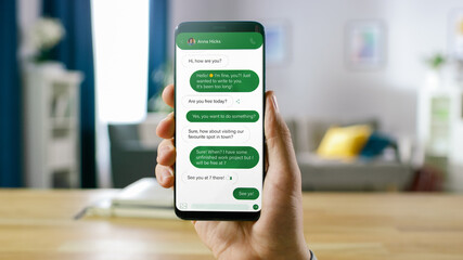 Close Up Shot of a Smartphone with Chat Messages About Date. Person is Holding Phone, Answering the Messages in Social Network. In the Background Cozy Homely Atmosphere.