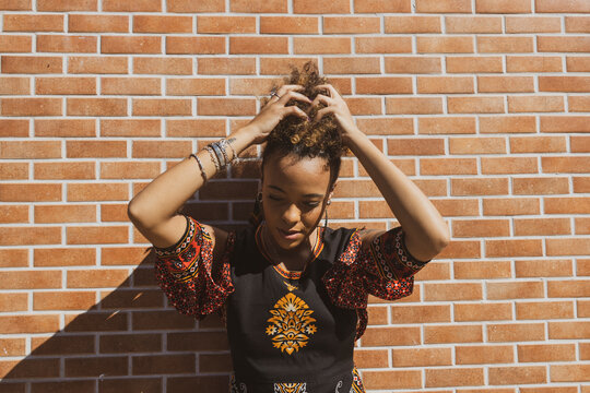 Beautiful African American woman wearing sunglasses leaning against  orange bricks wall tiding up her curly hair. Copy space for text.