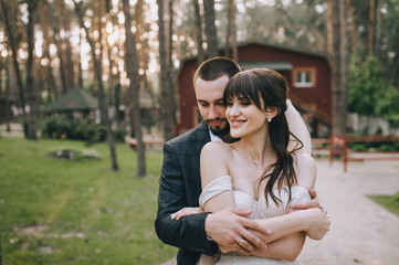 Beautiful newlyweds hug on the background of a villa, an estate in nature in the forest. wedding portrait of stylish groom in suit and cute brunette bride in white dress with veil.