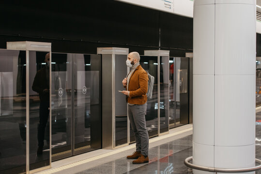 A full-length photo of a man in a face mask is holding a smartphone while waiting for a train at the subway platform. A bald guy in a surgical mask is keeping social distance.