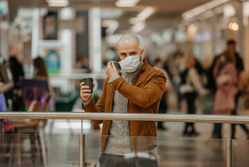 A man with a beard is putting on a medical face mask while holding a cup of coffee in the shopping center. A bald guy is keeping social distance.
