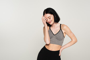 Smiling sportswoman with vitiligo standing with closed eyes isolated on grey