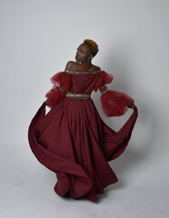 Full length portrait of pretty African woman wearing long red renaissance medieval fantasy gown, standing pose on a light grey studio background.