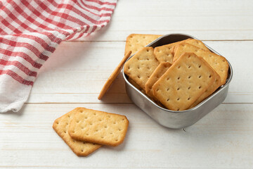 Cracker cookies in a stainless steel bowl with tablecloth on white wooden table background.