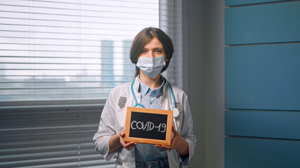 Fototapeta na wymiar Young woman doctor in white medical coat and face mask holds black covid 19 nameplate and looks straight