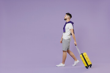Full size body length side view fun traveler tourist young brunet man 20s wear white t-shirt purple shirt go move stroll hold yellow suitcase bag isolated on pastel violet background studio portrait.