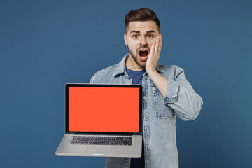 Shocked amazed wow young brunet man 20s wears denim jacket hold use work on laptop pc computer with blank screen workspace area touch cheek with hand isolated on dark blue background studio portrait