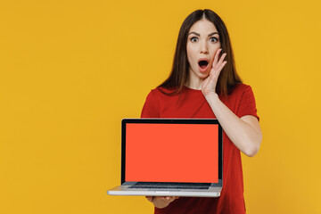 Naklejka premium Cute young brunette woman 20s wears basic red t-shirt hold use work on laptop pc computer with blank screen workspace area scream with hands near mouth isolated on yellow background studio portrait.
