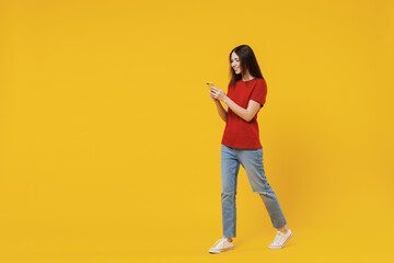 Full size body length fun satisfied young brunette woman 20s wears basic red t-shirt go move walk hold in hand use chatting searching mobile cell phone isolated on yellow background studio portrait