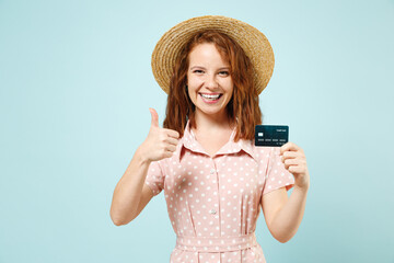 Smiling young redhead curly woman 20s hold in hand credit bank card showing thumb up like gesture wears casual pink dress straw hat look camera isolated on pastel blue color background studio portrait