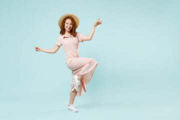 Full size body length happy smiling young redhead curly woman 20s dance spread hands raised leg wears casual pink dress straw hat look camera isolated on pastel blue color background studio portrait