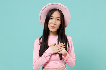 Charming smiling cute happy young brunette asian woman 20s wears pink clothes hat put folded hands on heart isolated on pastel blue color background studio portrait. People emotions lifestyle concept