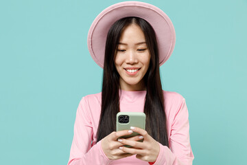 Pleased brunette asian woman 20s wear pink clothes hold in hand using mobile cell phone chatting browsing send sms to friend read news try to find information isolated on pastel blue color background