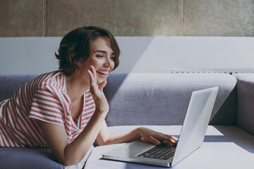 Young happy friendly woman 20s in casual clothes lying on grey sofa work online with laptop pc...