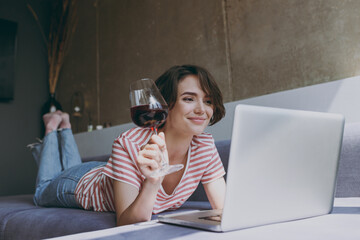 Full length young satisfied woman in casual clothes lying on grey sofa work online with laptop pc computer drink red wine hold glass rest relax indoors flat at home People lifestyle leisure concept.