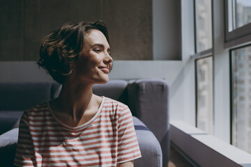 Close up young minded calm wistful caucasian woman 20s in casual clothes sitting near grey sofa...