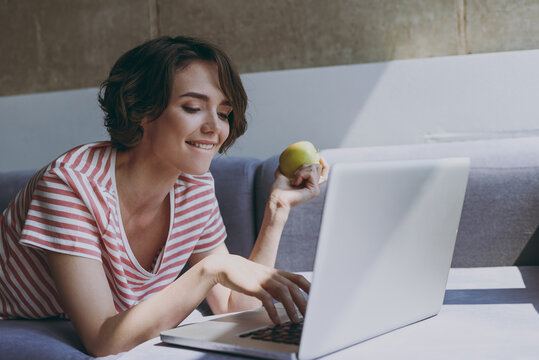 Young vegeterian woman in casual clothes lying on grey sofa work online with laptop pc computer typing message eat green apple fruit rest relax indoors flat at home People lifestyle leisure concept