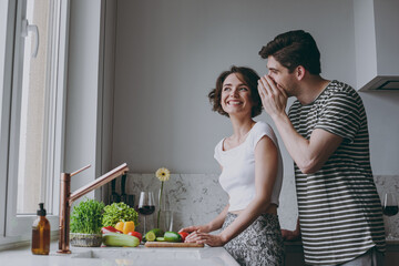 Young happy couple two woman man 20s in casual clothes boyfriend whisper gossip and tell secret behind his hand sharing news cook food in light kitchen at home together Healthy diet lifestyle concept