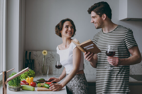 Young couple two woman man 20s in casual t-shirt clothes prepare vegetable salad read recipe book drink red wine cook food in light kitchen at home together. Healthy diet lifestyle vegeterian concept.