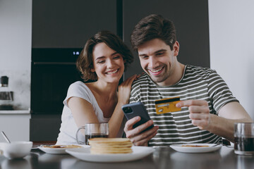 Young couple two woman man in casual clothes sit table eat pancake use mobile cell phone credit bank card shop online order delivery book tour in light kitchen at home together Healthy diet concept