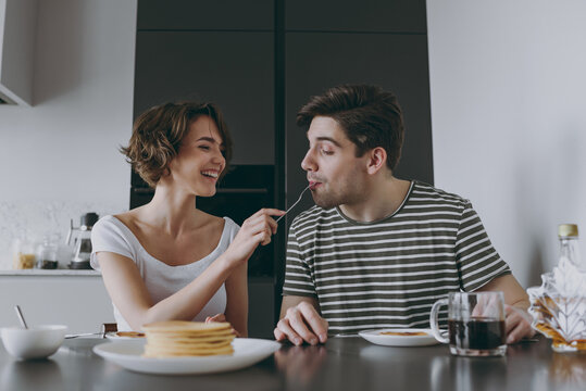 Young happy couple two woman man 20s in casual t-shirt clothes sit by table eat pancakes with maple syrup feed boyfriend cooking food in light kitchen at home together Healthy diet lifestyle concept.