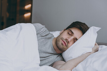 Calm brunet tranquil european young man 20s wearing pajamas grey t-shirt lying in bed sleep slumber...