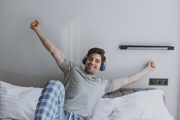 Fun happy satisfied young man wear grey t-shirt blue pajamas headphones sit in bed listen to music...