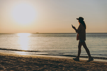 Side view full length young woman wearing straw hat shirt summer clothes walk use modile cell phone outdoors at sunrise sun dawn over sea beach background. People vacation lifestyle journey concept