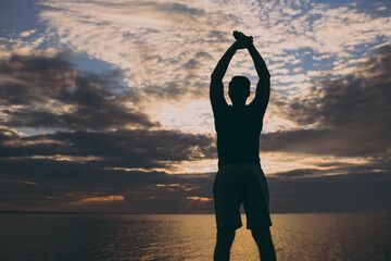 Back rear view young strong sporty fit sportsman man 20s wearing sports clothes warm up training do stretch exercise raise up hands at sunrise sun over sea beach outdoor seaside in summer day morning.