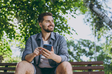 Bottom view smiling minded young man 20s in blue shirt sit on bench use mobile cell phone look aside rest relax in spring green city sunshine park outdoors on nature Urban lifestyle leisure concept