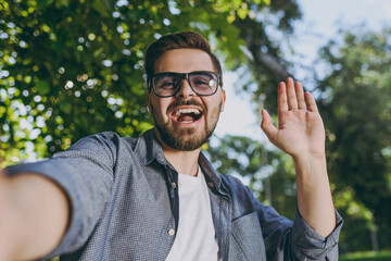 Close up young man 20s in blue shirt glasses do selfie shot on mobile cell phone show thumb up rest relax in spring forest garden green city park outdoors on nature. Urban lifestyle leisure concept.