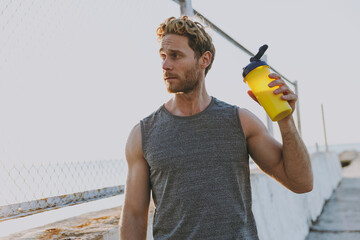 Young pensive sporty strong athletic fit sportsman man in sports clothes warm up train drink water hold bottle look aside at sunrise sun over sea beach outdoor on pier seaside in summer day morning © ViDi Studio