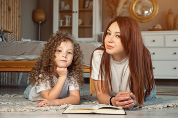 Smiling mom and preschooler daughter with book look at camera lying on floor at home, sunlight