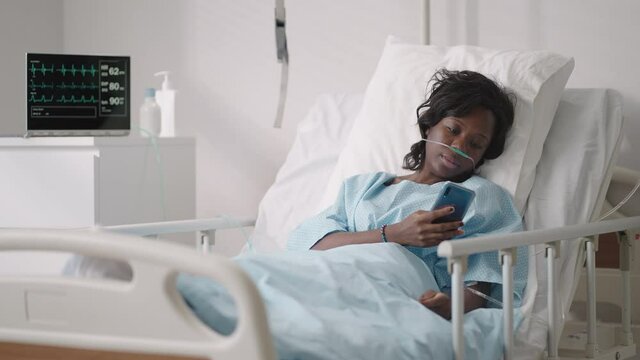 A young woman writes a message on her phone while lying in a hospital ward. An African girl is lying in a ward connected to ECG and oxygen devices in a mask and writes messages to relatives