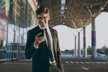 Traveler happy businessman young man 20s wear black dinner suit glasses standing outside at international airport terminal using mobile phone booking taxi order hotel Air flight business trip concept