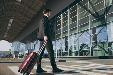 Full size side bottom view young traveler businessman man 20s in black dinner suit walk go outside at international airport terminal with suitcase valise crossing road Air flight business trip concept
