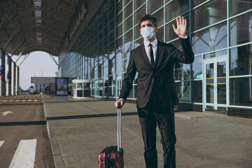Young traveler businessman man 20s wear black dinner suit sterile facial mask walking going outside at international airport terminal with suitcase valise waving hand Air flight business trip concept.