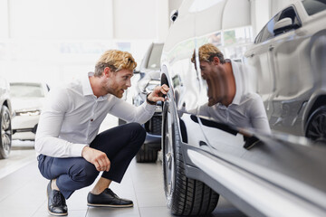 Obraz premium Full size man customer male buyer client in classic shirt chooses auto wants to buy new automobile touch car tire wheel disk in showroom vehicle salon dealership store motor show indoor Sales concept.