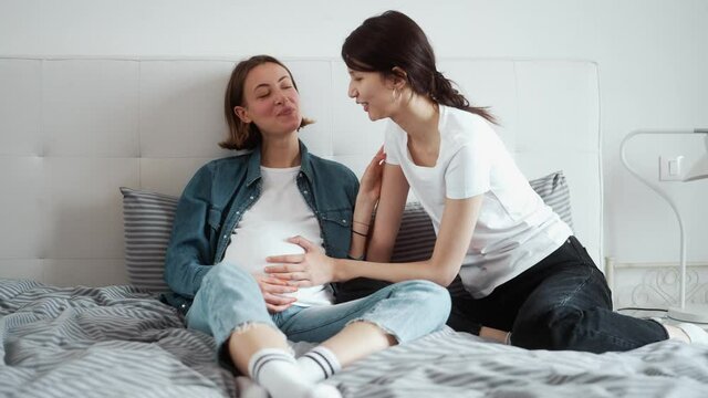 Happy lesbian girls expecting a baby and touching their belly while talking to each other sitting in the bedroom