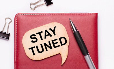 On a light background there are black paper clips, a pen, a burgundy notepad a wooden board with the text STAY TUNED